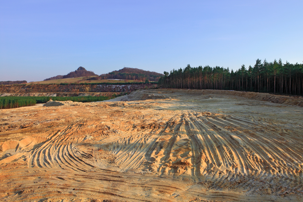A large cleared sandy area with tire tracks, bordered by a forest and distant hills under a clear sky.