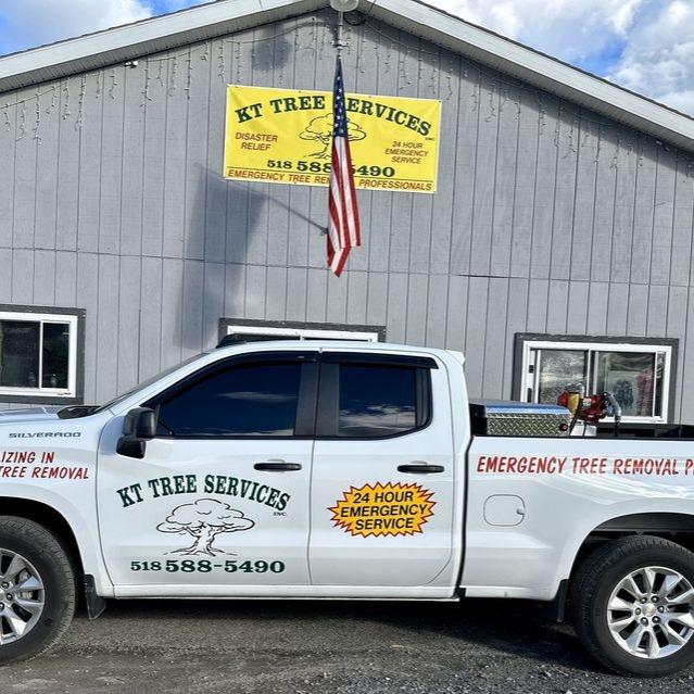 A KT Tree Services truck is parked in front of a gray building with a company sign and an American flag. The truck advertises 24-hour emergency tree removal service.